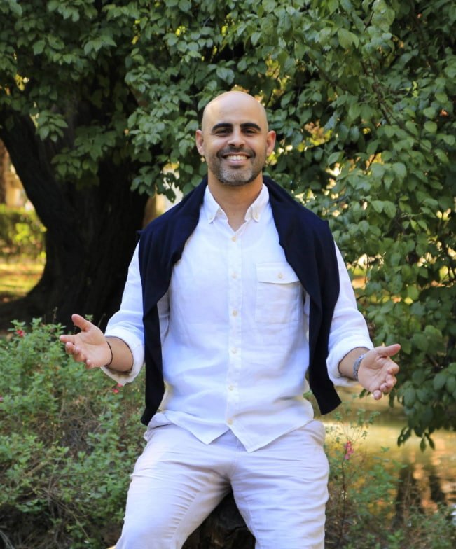 Coach sonriente con los brazos abiertos, de pie en una zona de árboles del Parque de Retiro, de frente a la cámara